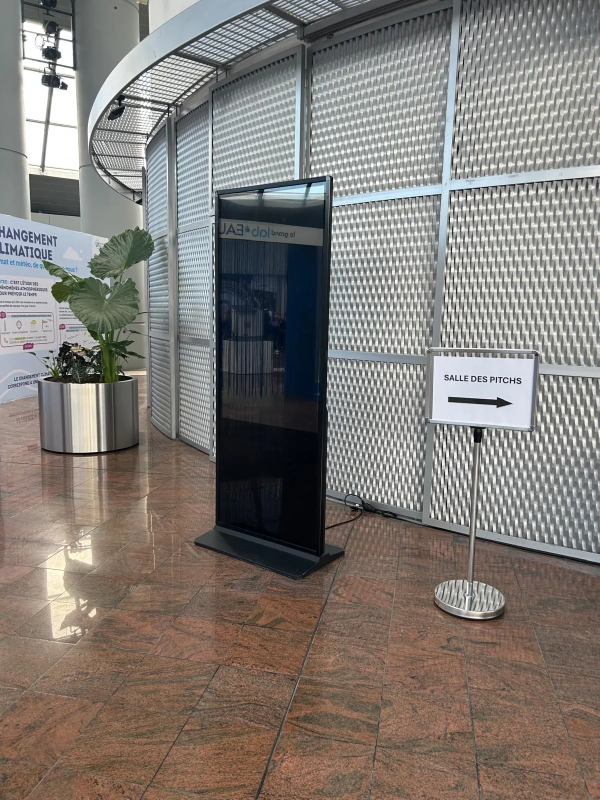 Totem numérique noir vertical installé dans le hall d'un centre de congrès à côté d'un panneau directionnel