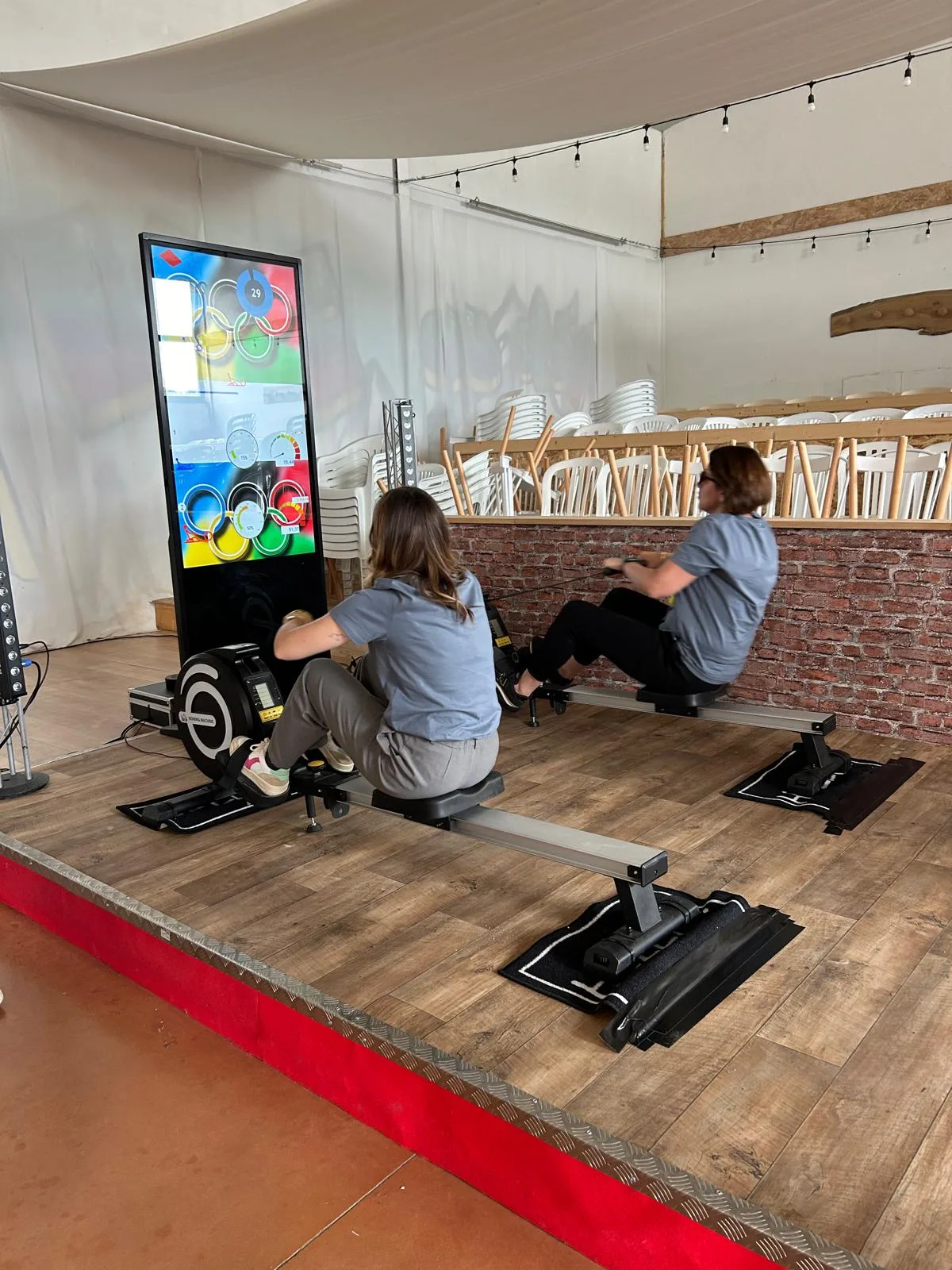 Deux participants s'affrontent sur des rameurs indoor face à un totem numérique dans une salle événementielle