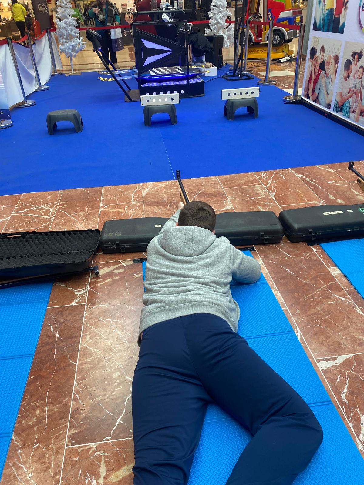 Homme en sweat gris allongé sur un tapis bleu visant trois cibles électroniques avec une carabine laser dans un centre commercial