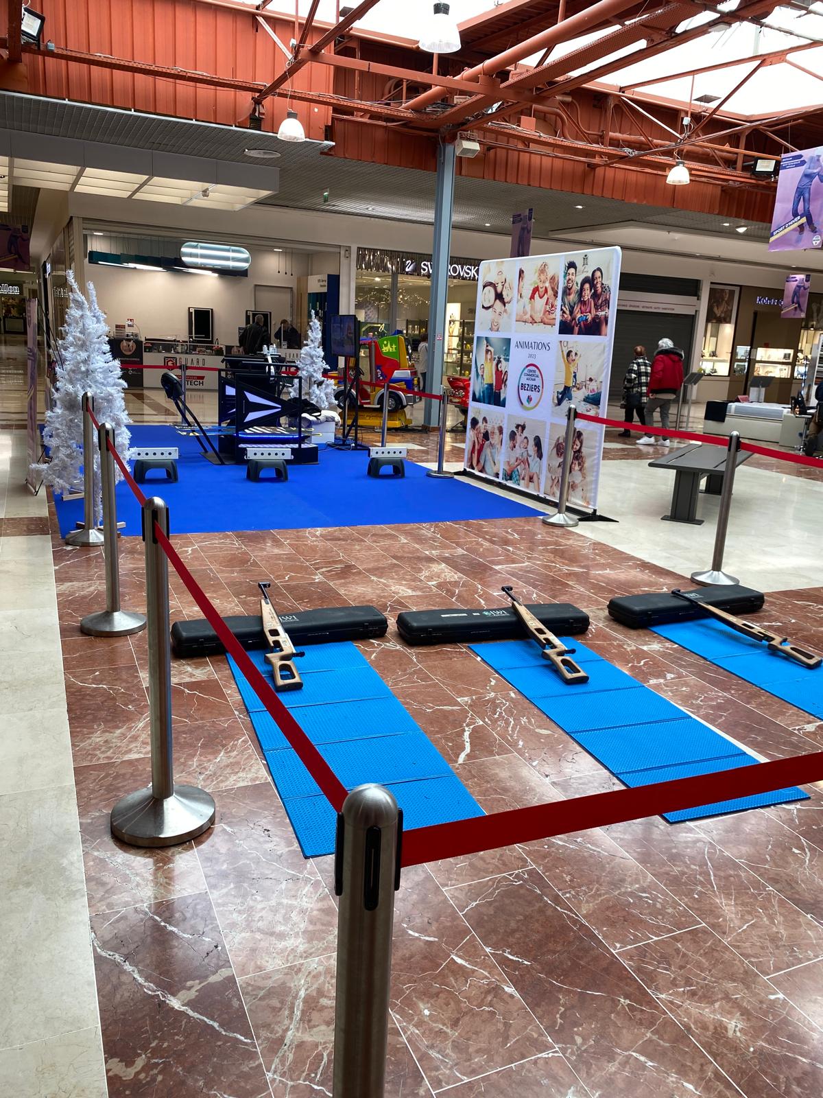 Stand de tir à la carabine laser avec trois postes sur tapis bleus et potelets à cordons rouges dans un centre commercial